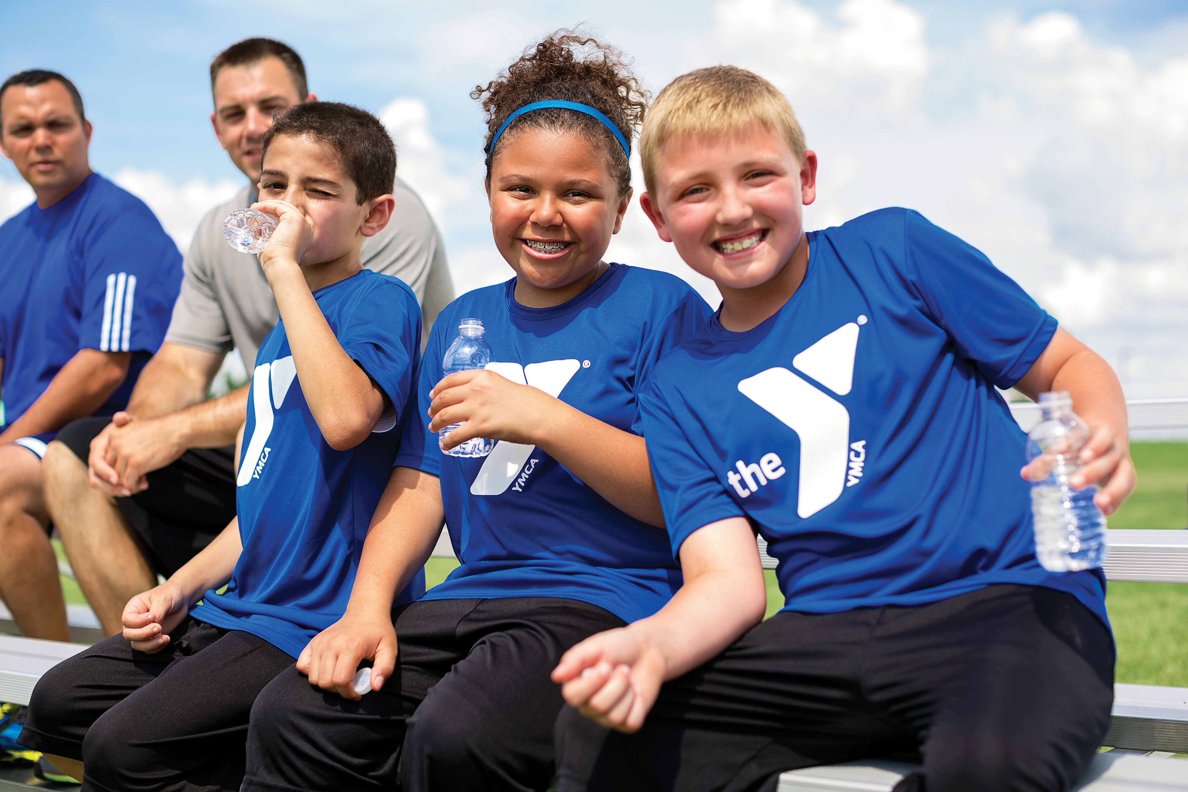 Three kids in blue YMCA tshirts smiling on the sidelines of a soccer game