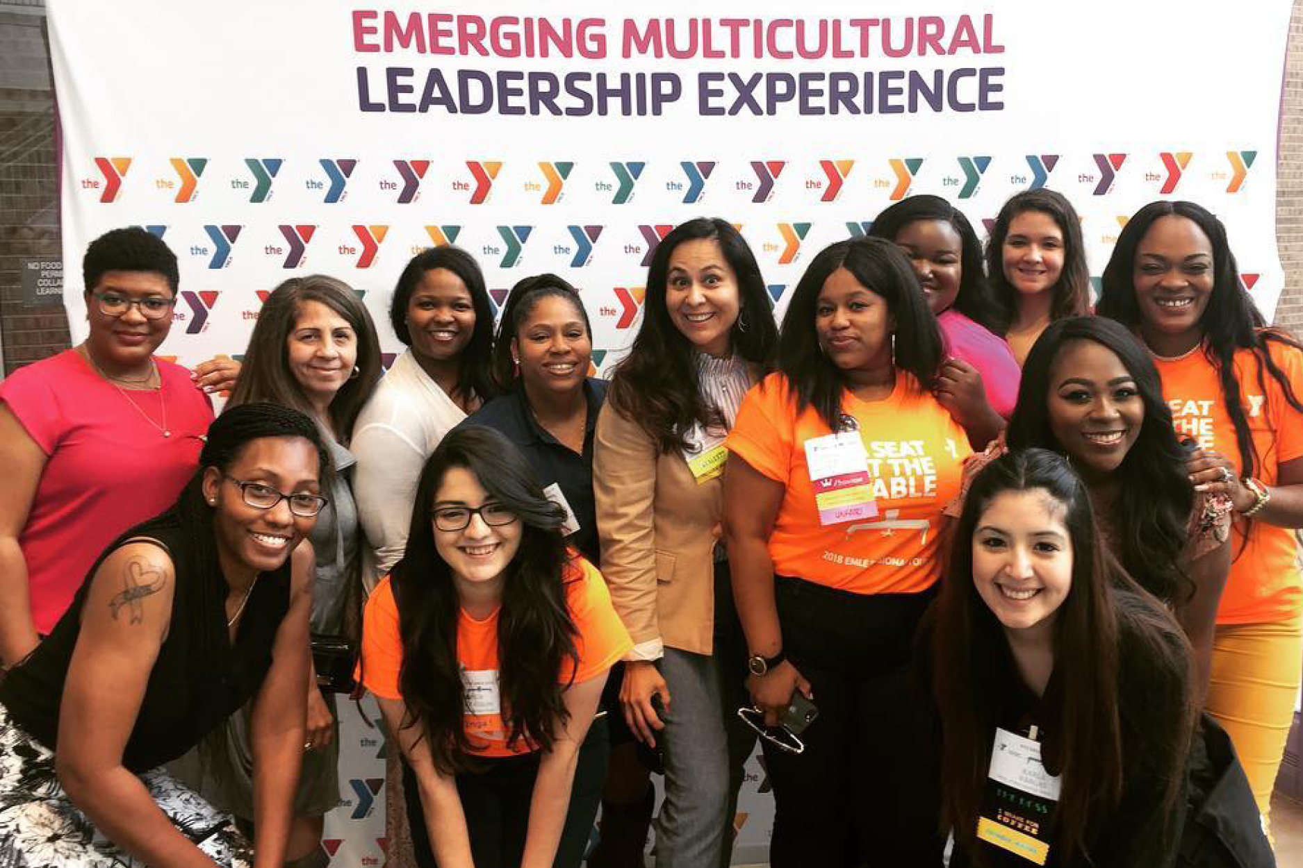 A group of multicultural women smiling and standing in front of a step-and-repeat reading "Emerging Multicultural Leadership Experience"