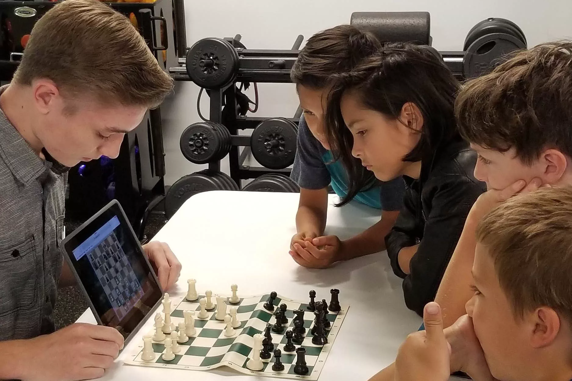 Four chess students gathered around instructor, chess board, and iPad, learning the rules of the game