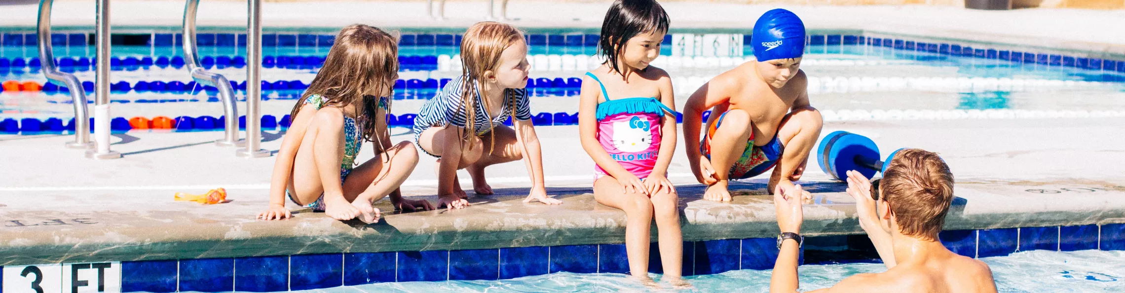 Four children crouching on edge of the pool, listening to lifeguard who is in the water